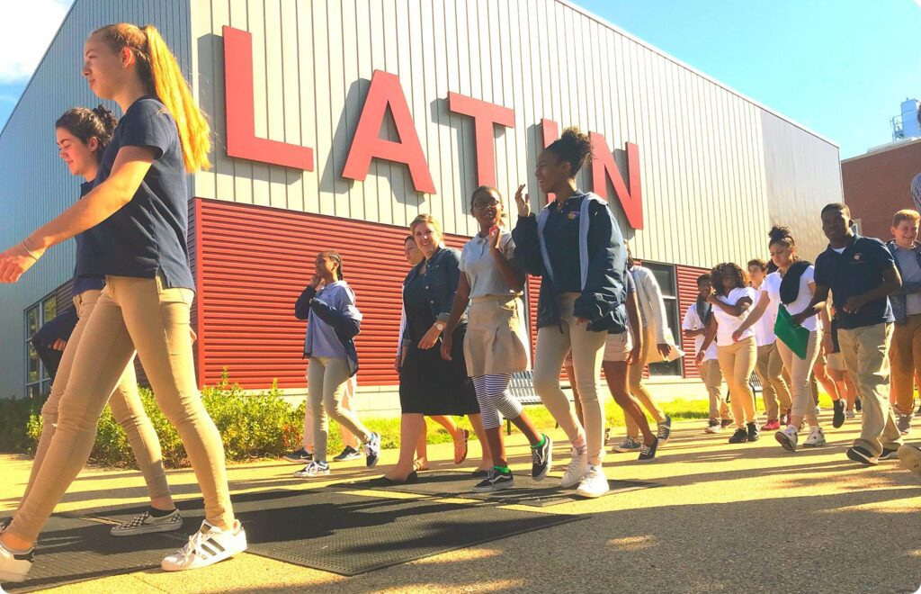 Students in uniform walk past a building with the word "LATIN" in large red letters on its side.